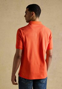 Homme de dos portant un polo à manches courtes orange vif et un jean bleu, sur un fond beige uni.