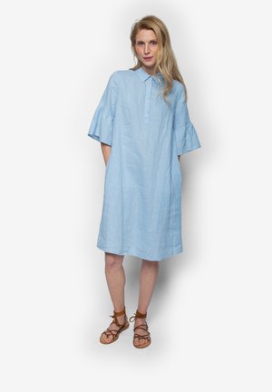 Blondynka stojąca w luźnej, jasnoniebieskiej sukience do kolan z krótkimi rękawami typu dzwonek i brązowych, wiązanych sandałach na płaskim obcasie.