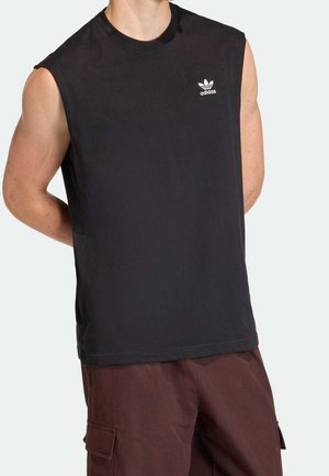 Mies, jolla on musta hihaton Adidas-paita, jossa rintalogo, ja ruskeat cargo-housut, kädet selän takana yksiväristä taustaa vasten.