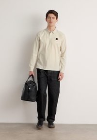 Beige langermet skjorte med brystlomme, kombinert med mørke teksturerte bukser. Modellen bærer en svart lærveske og har på seg grå sko.