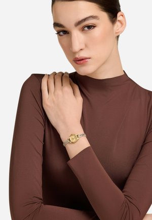 Goldene Armbanduhr mit runder Ziffernblatt und silbernem Gliederarmband, getragen an einer Hand, die auf einem braunen Langarmoberteil ruht. Einfaches Design, polierte Oberfläche.