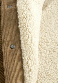 Tissu en velours côtelé marron avec boutons en métal à côté d'une doublure douce en sherpa de couleur crème, mettant en valeur des textures et matériaux contrastés.