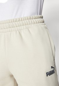 Lyse beige sweatpants med elastisk talje, der har et struktureret stof og et sort Puma-logo på venstre lår.