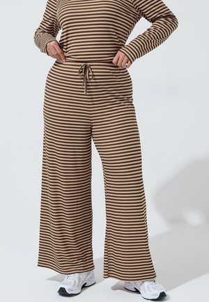 Person iført brune og beige bukser med brede ben og vandrette striber samt matchende langærmet top, iført hvide sneakers og med hænderne på hoften.