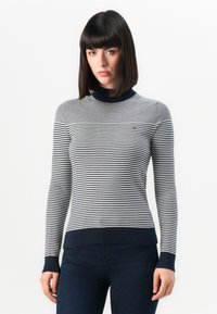 Stripet genser med høy hals, med marineblå og hvite horisontale striper, ribbede mansjetter og et hvitt emblem på brystet.