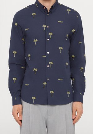 Chemise bleu marine à manches longues boutonnée avec des motifs de palmiers jaunes et le texte « DREAM », portée avec un pantalon gris clair.