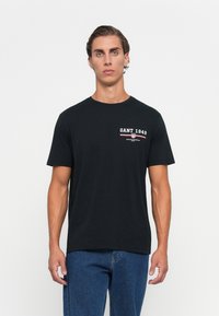 Ung mand med kort hår iført en sort GANT 1949 t-shirt og blå jeans, stående imod en simpel hvid baggrund.