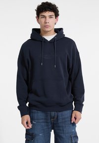 Sudadera con capucha de color azul marino en mezcla de algodón, con bolsillo frontal, cordón y logo en relieve. Corte holgado con puños de canalé.