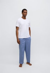 Hvit t-skjorte med korte ermer og en liten logo, og blå bukser med løs passform, stående barfot på en lys bakgrunn.