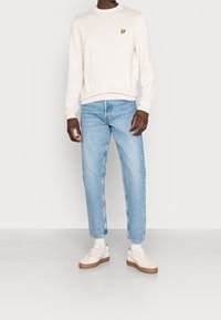 Sweatshirt beige clair avec un petit logo papillon jaune, associée à un jean droit bleu clair et des baskets blanc cassé avec des touches de beige.