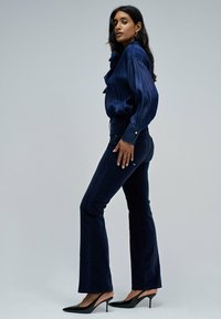 Mujer de pie de lado, vestida con una blusa de manga larga azul oscura, pantalones ajustados azul oscuro y zapatos de tacón alto negros con talón descubierto, sobre un fondo gris.