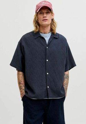 Jeune homme aux longs cheveux blonds portant une casquette rose, une chemise noire à manches courtes boutonnée sur un t-shirt bleu, avec les bras tatoués et les mains dans les poches.