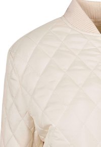 Chaqueta bomber acolchada de color beige claro con un cuello acanalado, que presenta un patrón de diamantes y una textura suave en todo el cuerpo.