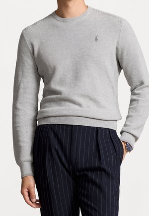 Mann trägt einen hellgrauen, strukturierten Pullover mit Rundhalsausschnitt und kleinem besticktem Logo, marineblaue, gestreifte, plissierte Hose und eine blaue Armbanduhr.