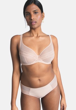 Frau trägt ein hellrosa BH mit Spitzenbesatz und passende Unterwäsche, steht vor einem schlichten hellen Hintergrund mit neutralem Gesichtsausdruck.