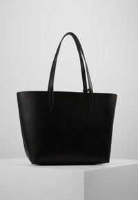 Černá kožená taška přes rameno s dvěma dlouhými uchy, strukturovaným tvarem, hladkým povrchem a minimalistickým designem. Žádné viditelné kovové doplňky nebo akcenty.