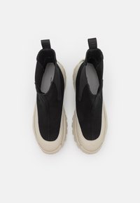 Zapatos negros sin cordones con parte superior texturizada, que presentan una suela de goma beige y una lengüeta. La tela suave contrasta con los acentos de goma.