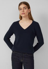 Granatowy sweter z dekoltem w kształcie litery V, o prążkowanej teksturze, z długimi rękawami i luźnym krojem. Noszony na ciemnych dżinsach, ukazujący proste, czyste linie.