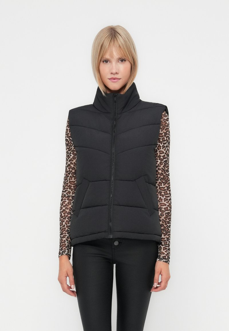 Zwarte gewatteerde vest met hoge kraag, ritssluiting, horizontale stiksels en twee voorzakken. Draagt over een lang shirt met dierenprint.