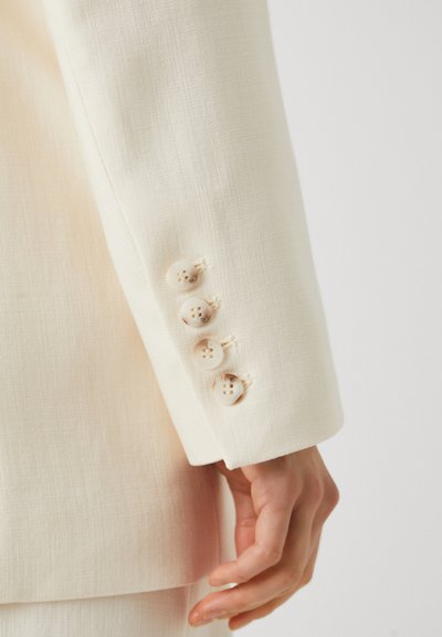 Manica di un blazer color crema con quattro bottoni chiari e un tessuto texturizzato. Il polsino presenta bordi puliti e dettagli di cucitura minimi.