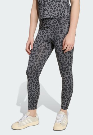 Femme portant des leggings gris et noirs à imprimé léopard et un haut assorti, debout, les mains relâchées le long du corps, portant des baskets blanches.
