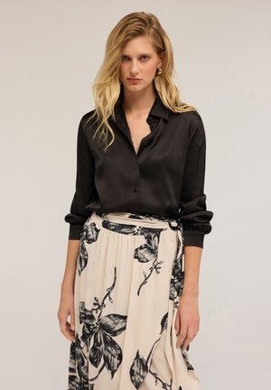 Donna con lunghi capelli biondi che indossa una blusa nera in satin con bottoni e una gonna bianca con grandi stampe floreali nere.