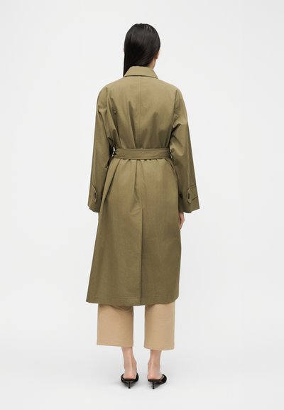 Γυναίκα με μακριά σκούρα μαλλιά φοράει χακί παλτό τύπου Trench με ζώνη, μπεζ παντελόνι μέχρι τον αστράγαλο και μαύρα παπούτσια με τακούνι, στέκεται με την πλάτη προς τα έξω.