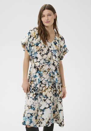 Kobieta z długimi brązowymi włosami ubrana w luźną sukienkę do kolan z abstrakcyjnym wzorem w kolorach niebieskim, czarnym i beżowym oraz krótkimi rękawkami.