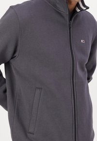 Grijze fleece jacket met ritssluiting en hoge kraag, voorzien van een klein logo, zijzakken en gestreepte binnenkraag. Zachte textuur.