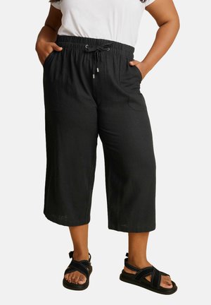 Pantalons noirs coupés avec une taille élastique, détail de cordon de serrage, poches latérales et tissu léger. Accompagnés de sandales noires.