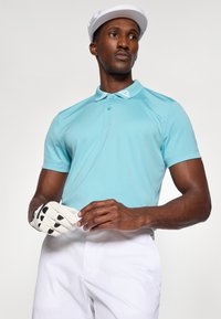 Lyseblå polo-skjorte med hvit krage og knapper, kombinert med hvite bukser. Modellen holder en hvit hanske med svarte detaljer.