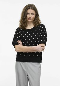 Melnas īsām piedurknēm adīta džemperis ar baltām punktiņām, ar apaļu kakla izgriezumu un brīvu piegriezumu. Kombinēts ar gaiši pelēkiem biksēm.