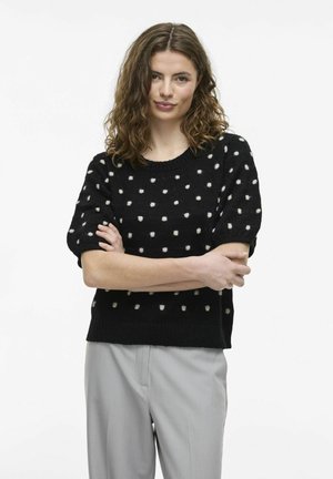 Suéter corto negro de mangas cortas con lunares blancos, textura de punto, cuello redondo y corte relajado. Combinado con pantalones gris claro.