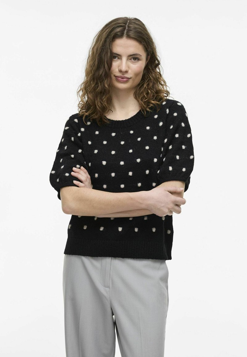 Melnas īsām piedurknēm adīta džemperis ar baltām punktiņām, ar apaļu kakla izgriezumu un brīvu piegriezumu. Kombinēts ar gaiši pelēkiem biksēm.