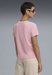 T-shirt in cotone rosa con maniche corte e scollo rotondo, mostrato da dietro. Pantaloni e accessori in tonalità della pelle aggiungono dettagli minimi.