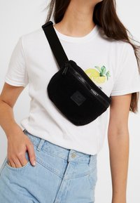 Czarna torba na ramię z polaru, z zamykaną na zamek kieszenią i fakturowanym wykończeniem, noszona na białej koszulce i zestawiona z jasnoniebieskimi dżinsami.