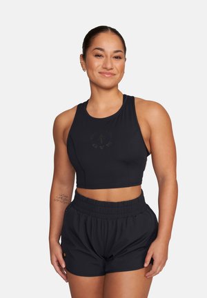 Smiling woman with hair tied back wearing black sleeveless crop top and black elastic-waist shorts, standing with hands relaxed.