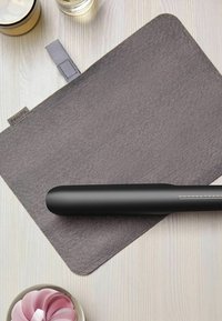 Lisseur à cheveux noir sur un tapis en feutre gris avec un bord arrondi, présentant une échelle de température sur le côté. L'arrière-plan comprend des textures douces.