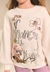 Svijetloružičasti duks s cvjetnim uzorcima i natpisom "Kindness" u crnoj boji, ukrašen šljokicama i nabranim rukavima.