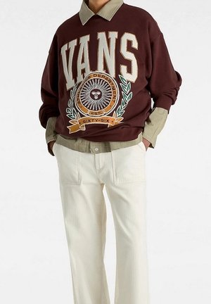 Personne portant un sweat-shirt Vans marron avec un logo graphique, superposé sur une chemise beige à col, associé à un pantalon blanc, mains dans les poches.