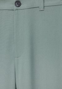 Primo piano di pantaloni verde chiaro che mostrano un bottone nero, la cintura, il passante per la cintura e i dettagli della tasca cucita.