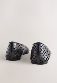 Paire de chaussures plates marron foncé avec motif découpé géométrique, vues de dos sur un fond clair neutre.