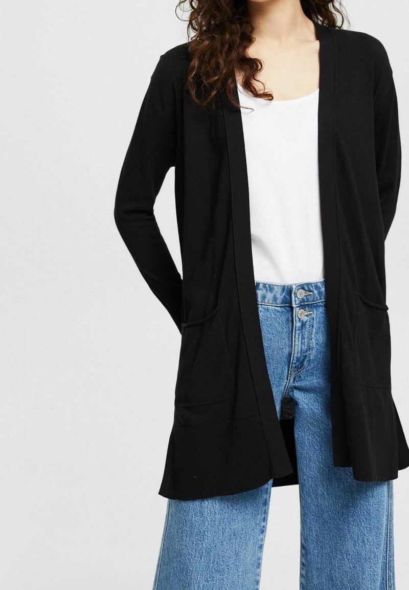 Femme portant un cardigan noir ouvert sur une chemise blanche et un jean bleu taille haute avec deux boutons, debout devant un fond uni.