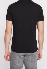 Homme portant un polo à manches courtes noir et un jean gris, vu de dos, sur fond blanc.