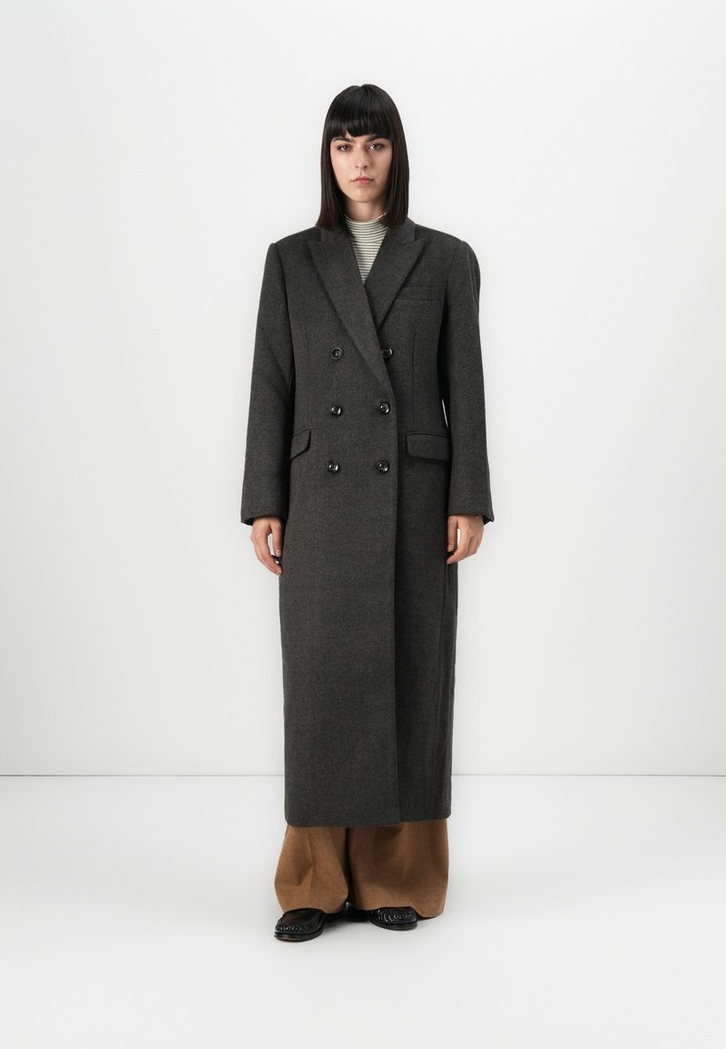 Femme aux cheveux noirs raides portant un long manteau croisé gris foncé sur un pantalon large marron et des chaussures noires, sur fond blanc.