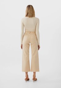 Femme se tenant de dos, portant un haut à manches longues en ribbed beige, un pantalon large cropped taille haute et des sandales à talons beiges.