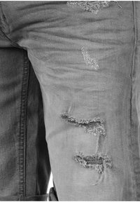 Jean en denim gris clair avec des détails usés, comprenant des bords effilochés, des déchirures et une texture lisse. Gros plan sur la partie gauche de la jambe.