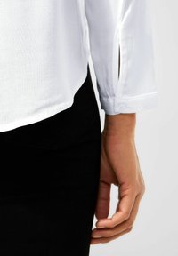 Camicia bianca a maniche lunghe con dettagli sui polsini a bottone, abbinata a pantaloni neri. Tessuto liscio, taglio dritto con spacco laterale sulle maniche.