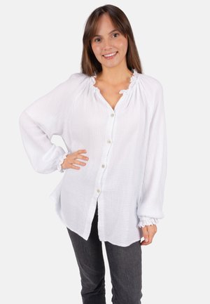 Jeune femme souriante, portant un chemisier blanc ample à boutons avec col et poignets à volants, accompagné d'un pantalon gris foncé, debout avec une main sur la hanche.