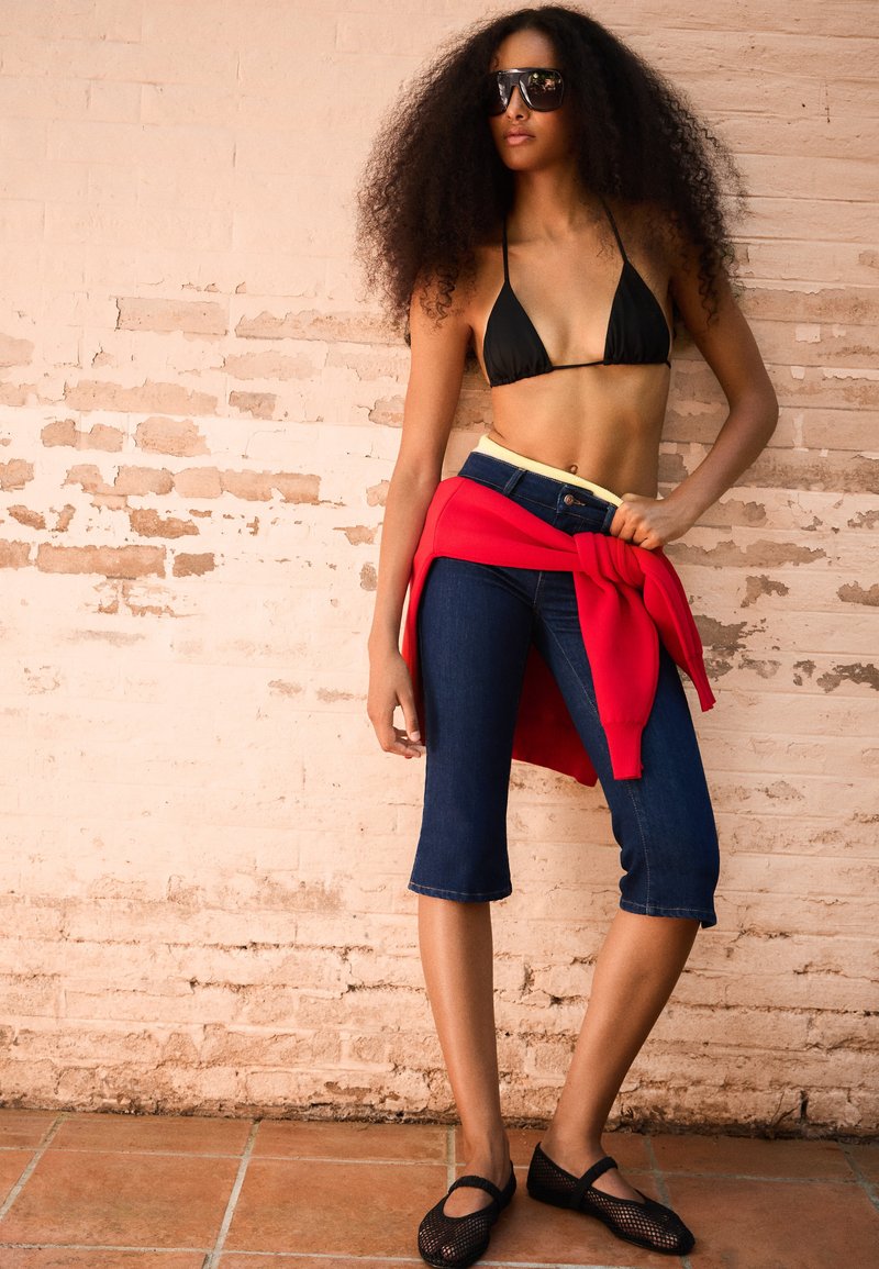 Femme aux cheveux bouclés portant un haut de bikini noir, un jean capri foncé, des ballerines en mesh noires, des lunettes de soleil et un sweat-shirt rouge noué autour de la taille contre un mur de briques.